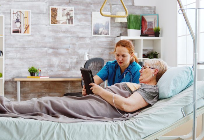 Female caregiver helps an old disabled woman lying in hospital bed to use a digital tablet PC. Bright room with big windows